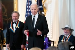 El presidente Joe Biden con los homenajeados del Centro Kennedy 2023, Billy Crystal, izquierda, y Barry Gibb, derecha, en el podio del salón Este durante una ceremonia en honor a los homenajeados del Centro Kennedy en la Casa Blanca, el domingo 3 de diciembre de 2023.