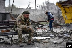 Seorang prajurit Ukraina merokok sambil duduk di bangku ketika seorang penduduk setempat membersihkan puing-puing di dekat sebuah bangunan yang rusak akibat serangan udara Rusia di Kota Orikhiv, wilayah Zaporizhzhia, Ukraina, 5 April 2024. (Foto: AP)