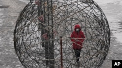 一位身著紅衣戴著口罩的北京婦女正走過一個球形的編制藝術作品。中國實行的新冠病毒”清零“政策名為保護人民和經濟，但實際效果可能會使中國更難擺脫疫情的肆虐。（2022年1月22日）