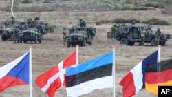 FILE - Flags are seen in front of soldiers with military vehicles during NATO exercises near Swietoszow Zagan, Poland, June 18, 2015. Russia is demanding that the alliance reduce its size in Europe to levels it maintained in 2000.