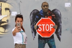 A woman takes a selfie picture posing next to a street poster artwork by Italian urban artist Salvatore Benintende aka "TVBOY" depicting George Floyd with angel wings and holding a stop traffic sign against racism, in a street of Barcelona