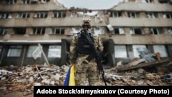 图库照片：一名手持乌克兰国旗的乌克兰女军人，背景是座被炸毁的房屋。(来源：Adobe Stock)