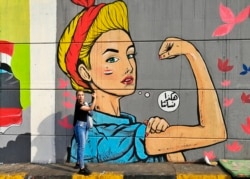 In this Monday, Nov. 18, 2019 photo, a woman poses for a photo in front of graffiti at the Saadoun tunnel, in Baghdad, Iraq. The tunnel that passes under Baghdad's landmark Tahrir Square has become an ad hoc museum for Iraq's revolution: Young artists dra