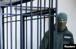 FILE - A member of the National Anti-Corruption Bureau of Ukraine stands guard next to a defendant's cage during a court hearing in Kyiv, April 21, 2017.
