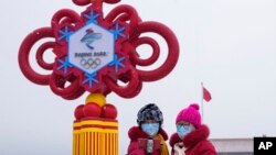 Dos mujeres, con mascarillas con el eslogan de los JJOO de Invierno de Beijing, se toman un selfie con una pieza decorativa instalada en la Plaza de Tiananmen con motivos de los Juegos, en Beijing, el 20 de enero de 2022. 