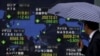 Seorang pejalan kaki yang memegang payung melihat papan elektronik indeks pasar saham berbagai negara di Tokyo, Jepang, 6 Juli 2015. (Foto: Reuters)