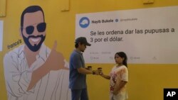 Una pareja se encuentra frente a un mural político del presidente Nayib Bukele, el mensaje: "Ordeno que vendan 3 pupusas por un dólar", parte de una ofensiva del gobierno para bajar los precios de los alimentos, en San Salvador, El Salvador, el viernes 12 de julio de 2024.
